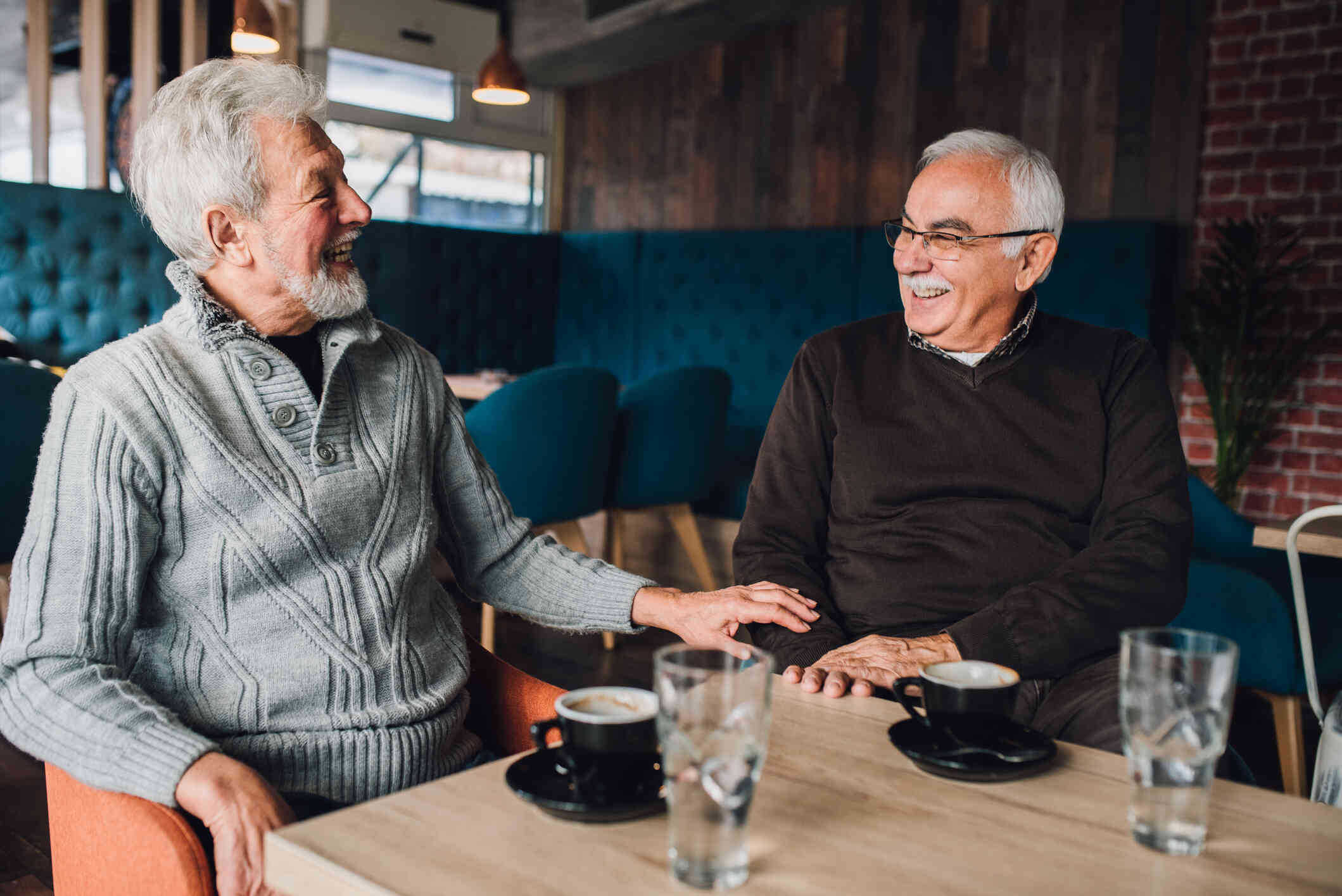 Two mature men in sweaters smile at each other as they sit at a table in a cafe with cups of coffee and water in front of them.